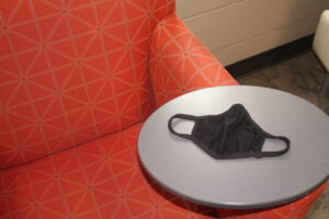 A black, cloth mask sits on a table with a chair in the background.