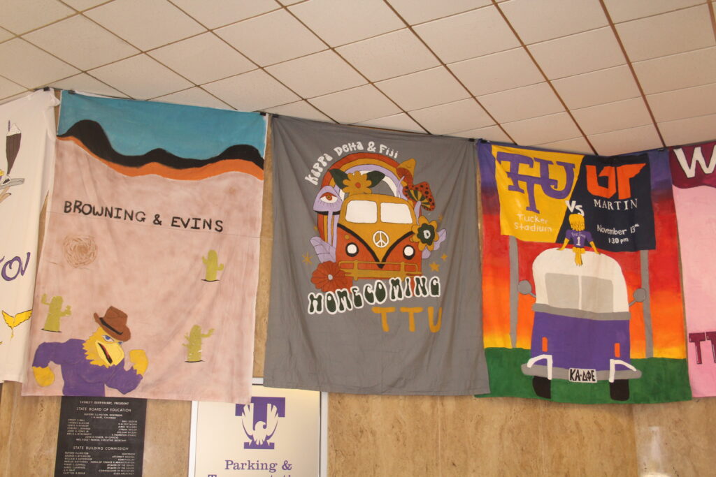Banners hanging in RUC with Awesome Eagle wearing a cowboy hat, a Volkswagen Microbus, and people sitting on top of a bus looking at a scoreboard.