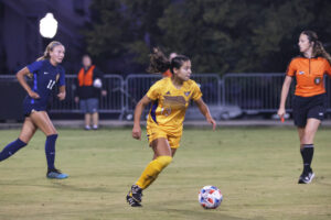 A Tech soccer player advances the ball with a player and referee in the background.