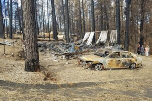 A completely burnt car sits in front of the foundation of burned cabin.